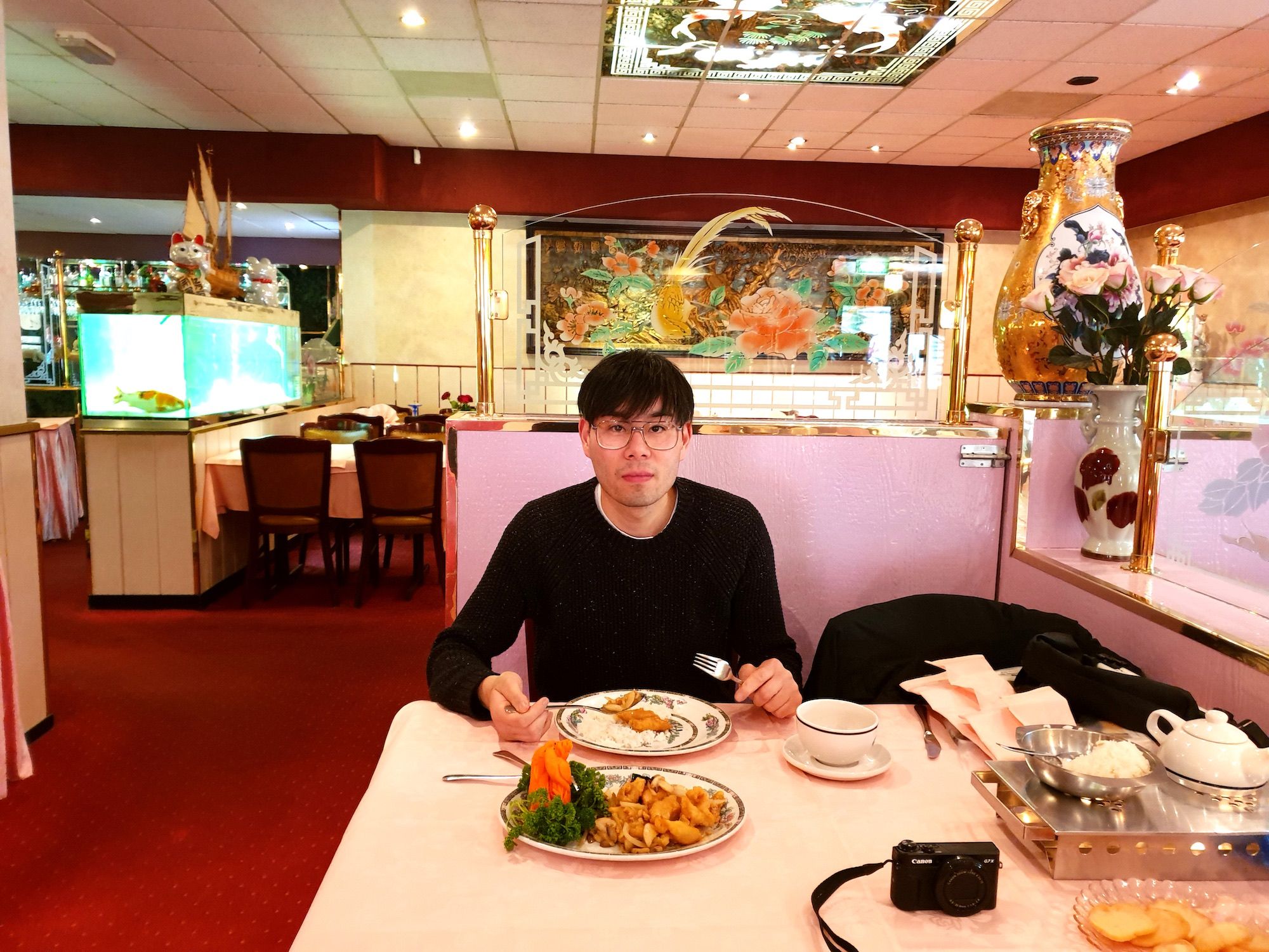 Portrait of the artist Benjamin Li – black hair, black sweater and glasses – sitting in the 'Chinees-Indisch' Restaurant Peach Garden, eating dinner.