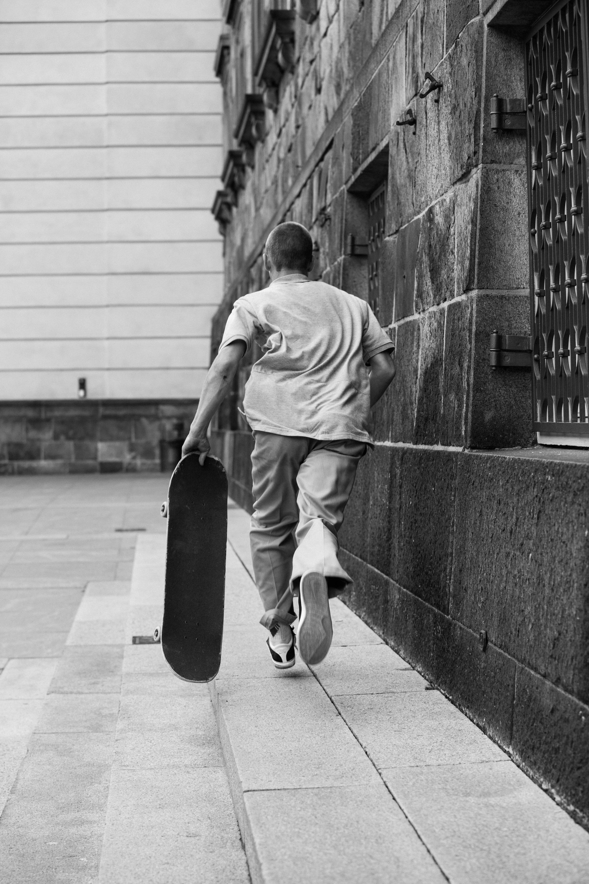 A black and white image of the back of a young man running down the street with a skateboard.