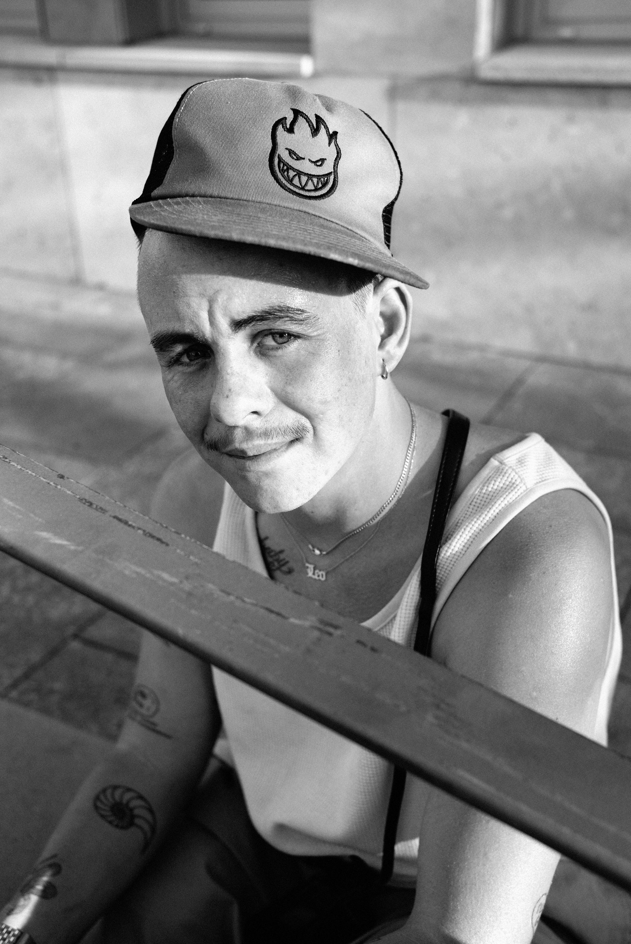 Black and white portrait of a man looking at the camera wearing a white tank, sideways Spitfire cap and chain necklaces.