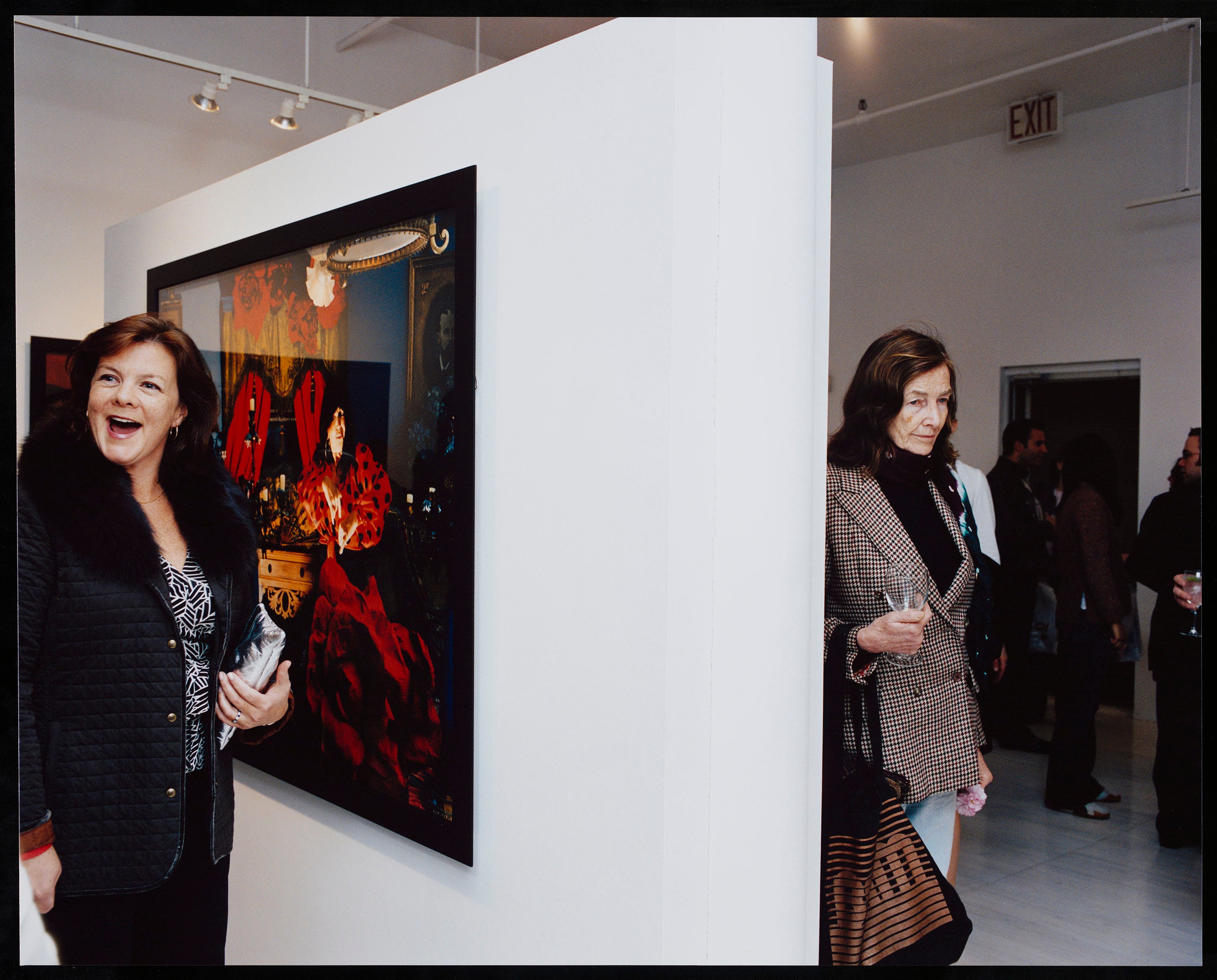 Two ladies are standing in an opening on two sides of a wall. One lady smiles and looks to the left, the other lady looks down and does not smile. In the background white walls with works of art and other visitors.