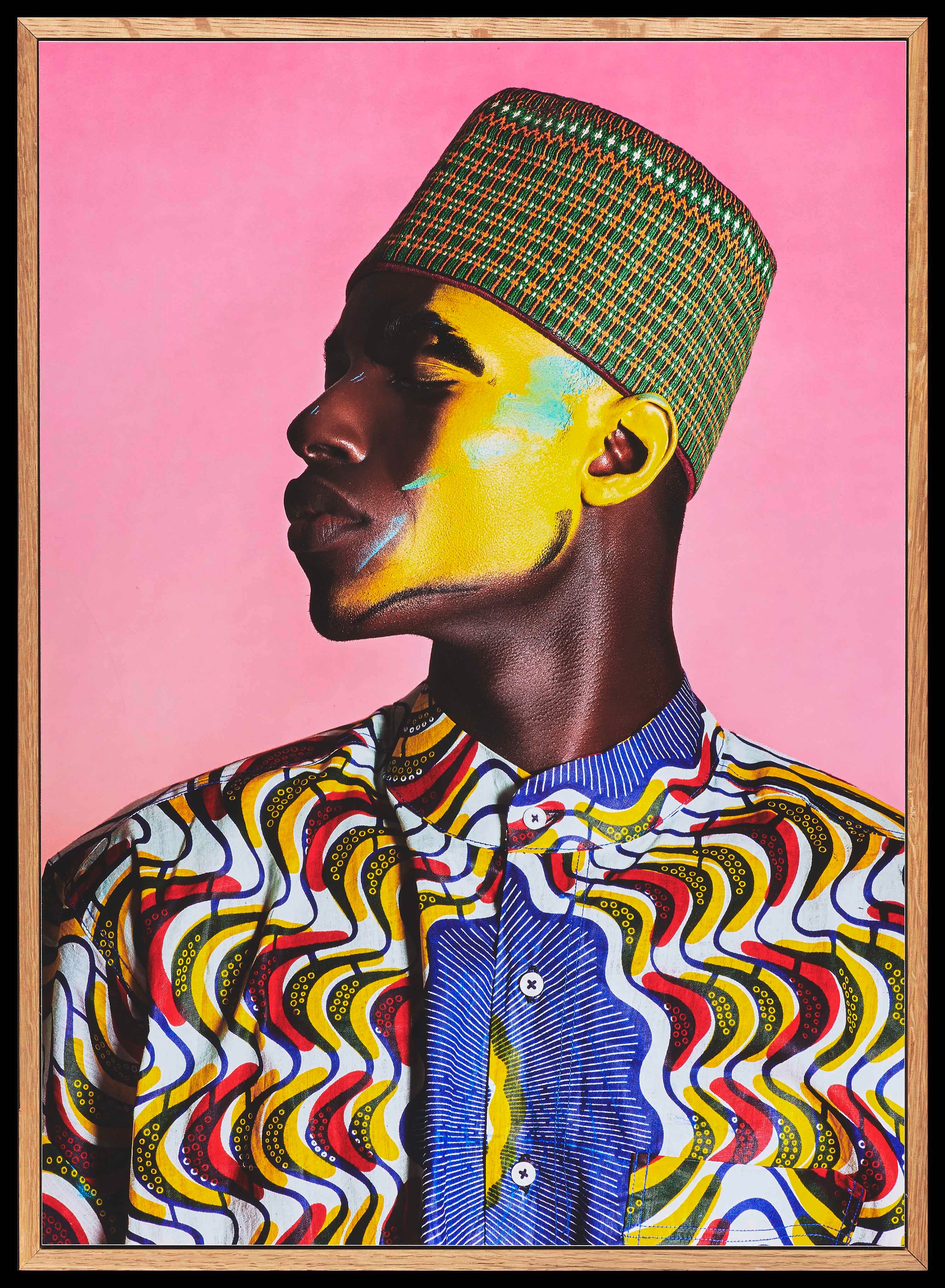 Portret van de zijkant van een deels geel geverfd gezicht van een man met een groen hoedje op, gekleed in kleding met Afrikaanse tribal-print, tegen een roze achtergrond.