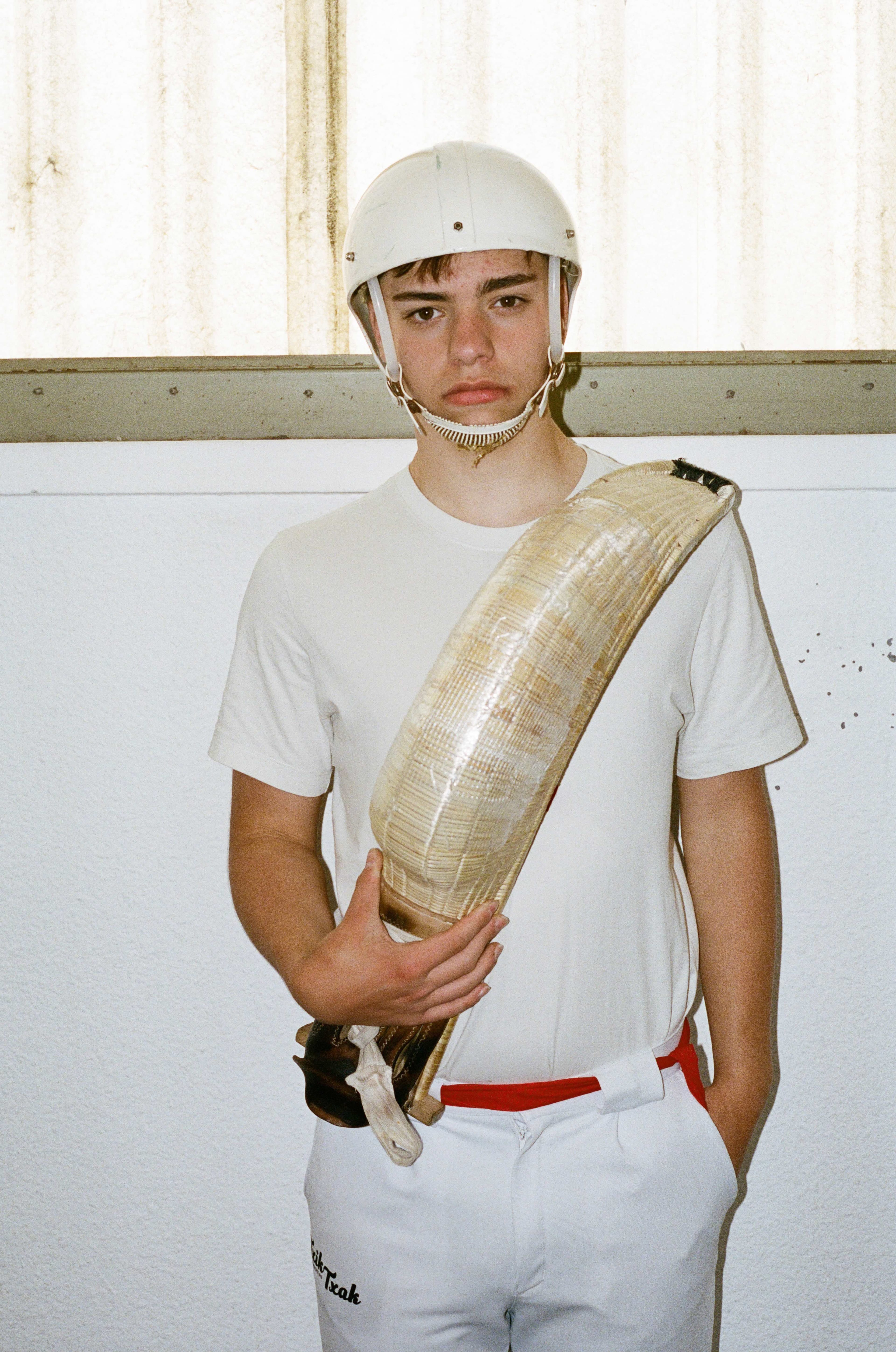 A young man in a white sports uniform and helmet holds a jai alai cesta across his torso.