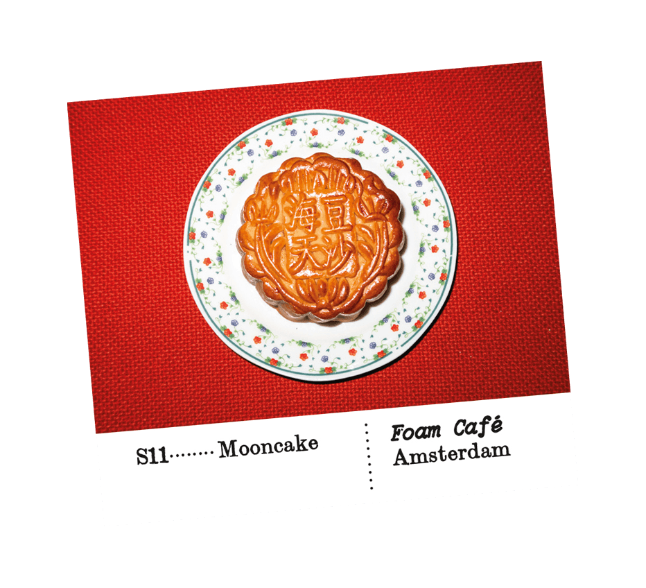 Image of a sticker, showing an image of moon cake on a plate, on a red background, with the subtitle: S11- Mooncake. Foam Café. Amsterdam. From the series In Search of Perfect Orange by artist Benjamin Li.